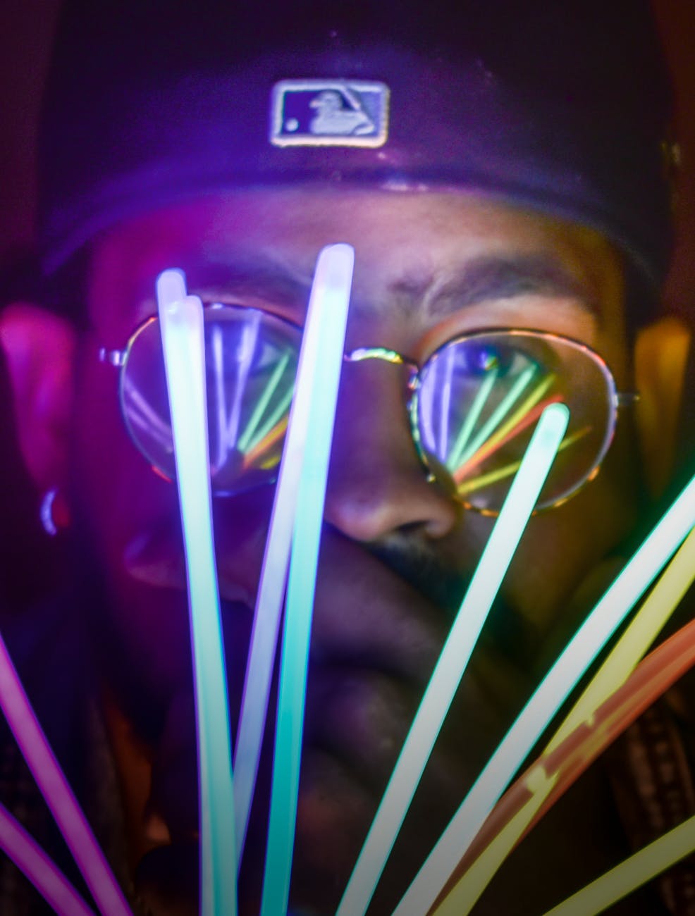 A dark-skinned gentleman with a backwards cap and glasses holds several glowing sticks in front of his face, lighting it up